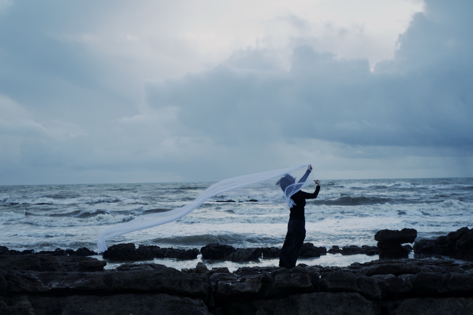 Dance with the Wind, distant figure extending fabric into the wind beside the sea