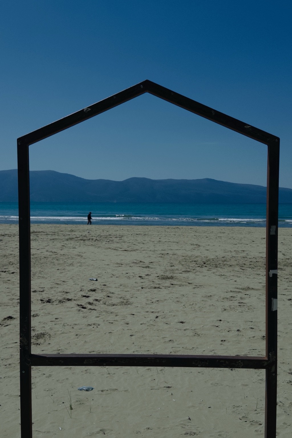 Gaze, house-shaped metal frame enclosing a distant walking figure on a beach