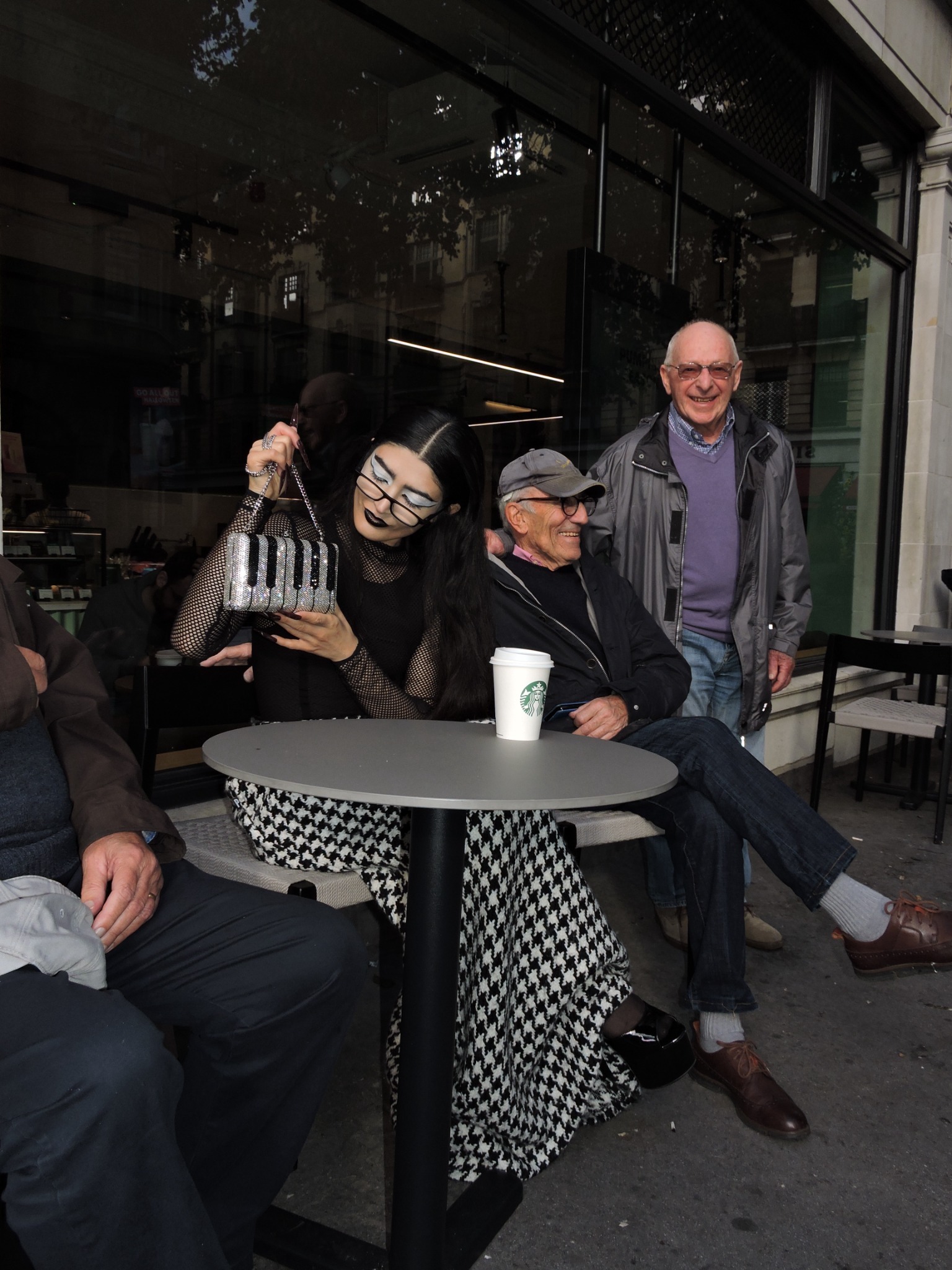 Her Spell, styled figure handling a piano-key handbag beside people at an outdoor table in London